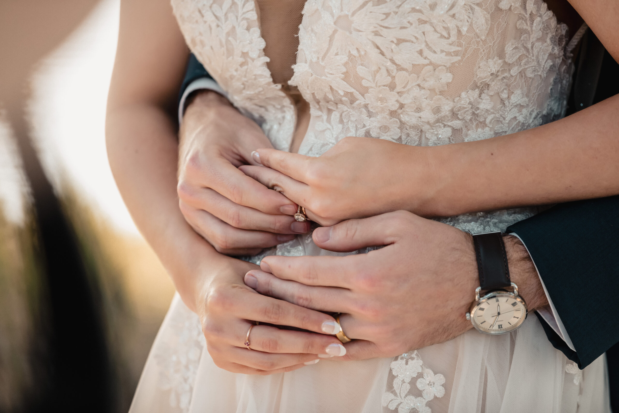 Close-up of a couple embracing, showing their hands intertwined. The bride is wearing a lace wedding dress, and the groom is wearing a watch and a dark suit